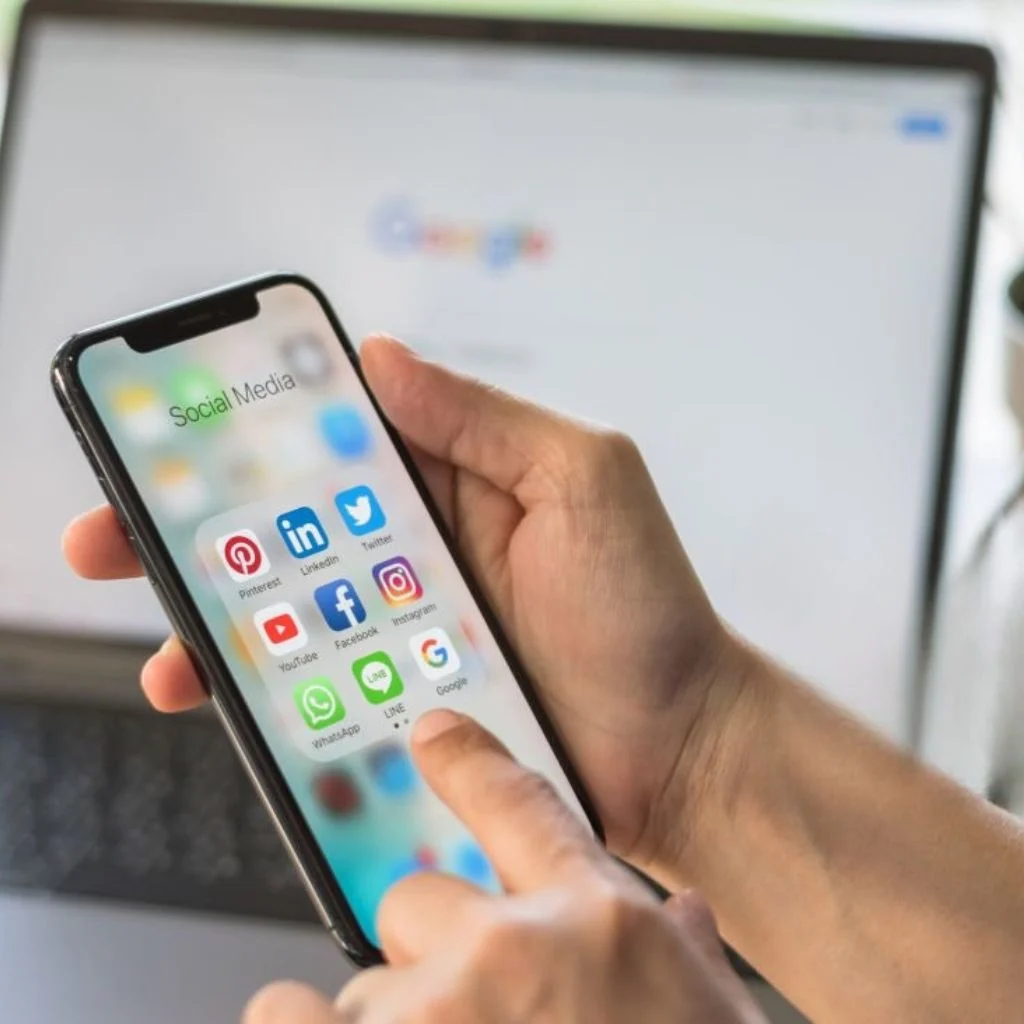 Hands holding a smartphone with social media apps and a laptop showing digital search in the background